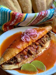 A mexican sandwich called torta ahogada, with carnitas and refried beans inside a crusty birote salado, drowned in a spicy tomato salsa. topped with pickled red onions with some lime wedges on the side.