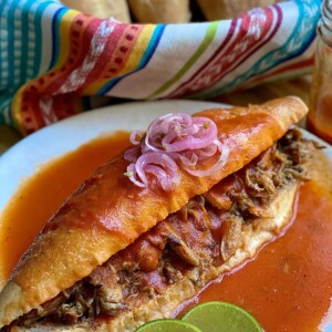 A mexican sandwich called torta ahogada, with carnitas and refried beans inside a crusty birote salado, drowned in a spicy tomato salsa. topped with pickled red onions with some lime wedges on the side.