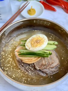 A bowl of Korean mul naengmyeon (cold noodles) features slices of beef, cucumber strips, and half a boiled egg on top. Garnished with sesame seeds, it's served in a stainless steel bowl with chopsticks and mustard on the side.