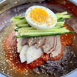 A bowl of chilled Korean noodles, known as bibim naengmyeon, topped with slices of beef, radish, julienned cucumber, a halved boiled egg, and sesame seeds, all served in a savory broth with a red chili sauce.