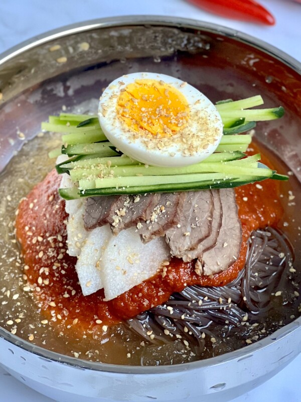 A bowl of chilled Korean noodles, known as bibim naengmyeon, topped with slices of beef, radish, julienned cucumber, a halved boiled egg, and sesame seeds, all served in a savory broth with a red chili sauce.