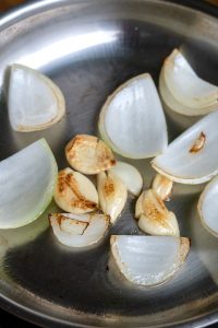 Sliced onions and garlic cloves are being sautéed in a stainless steel pan, with some pieces lightly browned, reminiscent of flavors in al pastor cooking.