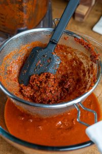 A black spatula presses an al pastor-inspired, red chunky mixture against a fine-mesh strainer, letting smooth sauce drip into a clear bowl below. The setup rests on a wooden surface, with a blender containing more of the savory mixture in the background.