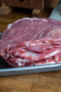 A stack of raw, marbled beef slices, reminiscent of al pastor preparation, sits on a metal tray placed on a wooden surface. The meat appears fresh with visible fat layers, ready for preparation or cooking.