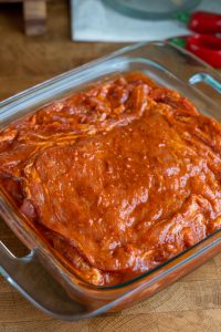 A glass dish contains raw al pastor meat marinated in a thick, red sauce. The marinade appears spicy, with a rich, glossy texture. Red chili peppers and a wooden surface are visible in the background.