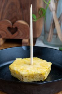 A slice of pineapple with a long wooden skewer, reminiscent of an al pastor setup, is placed upright in a black pan. A wooden board with a heart cut-out and a glass jar filled with wooden utensils enhance the background ambiance.