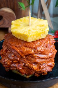 A stack of uncooked, marinated al pastor meat piled in layers on a vertical skewer, topped with a slice of fresh pineapple. The meat is heavily seasoned with a red sauce, displayed on a black plate. Wooden kitchen decor is in the background.