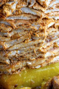 Close-up of layered, seasoned al pastor meat with a golden-brown crust, stacked tightly, suggesting it might be part of a shawarma or similar dish. A slice of yellow, juicy pineapple is visible at the bottom, adding a fresh contrast.