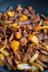 A close-up of strips of pork, pineapple chunks, and sliced onions cooking in a pan. The ingredients are browned and seasoned al pastor style, creating a savory and colorful dish reminiscent of traditional Mexican flavors.