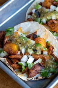 A close-up reveals tacos al pastor, generously topped with diced onions, pineapple, cilantro, and green salsa, all elegantly served on corn tortillas arranged in a metal tray.