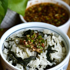 A bowl of Korean thistle cooked with rice and topped with a seasoning sauce containing sesame seeds and chopped green onions. In the background, a bowl of the seasoning sauce. A green plant is partially visible to the side.