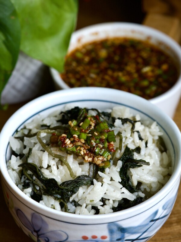 A bowl of Korean thistle cooked with rice and topped with a seasoning sauce containing sesame seeds and chopped green onions. In the background, a bowl of the seasoning sauce. A green plant is partially visible to the side.