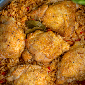 A close-up of a pan filled with seasoned chicken thighs atop spicy rice, reminiscent of Cuban arroz con pollo. The dish is garnished with bay leaves and surrounded by green leaves, making the freshly cooked food look incredibly flavorful.