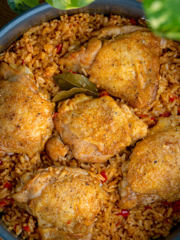 A close-up of a pan filled with seasoned chicken thighs atop spicy rice, reminiscent of Cuban arroz con pollo. The dish is garnished with bay leaves and surrounded by green leaves, making the freshly cooked food look incredibly flavorful.