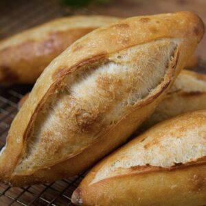 Freshly baked, golden-brown loaves of bread (called birote salado) are nestled on a cooling rack. The bread has a crunchy crust and is surrounded by green foliage in the background, adding a touch of natural decor.