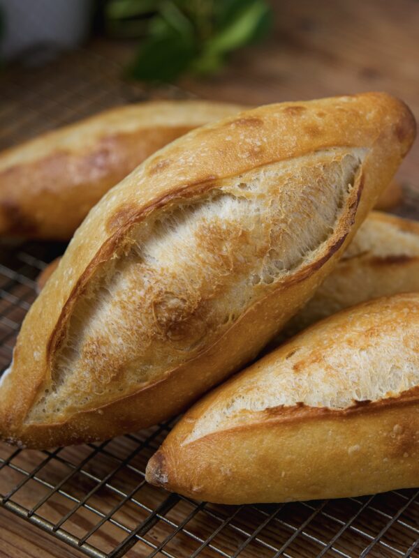 Freshly baked, golden-brown loaves of bread (called birote salado) are nestled on a cooling rack. The bread has a crunchy crust and is surrounded by green foliage in the background, adding a touch of natural decor.