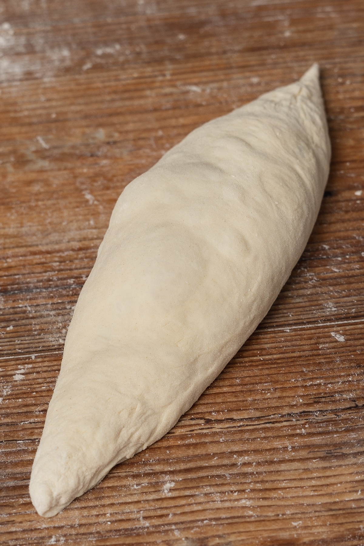 A piece of shaped bread dough rests on a wooden surface, ready for baking. The dough is elongated with tapered ends, resembling a boat shape. Dustings of flour are visible on the surface, enhancing the rustic appearance.