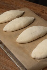 Four uncooked, elongated dough pieces rest on a parchment-lined inverted baking sheet. The dough appears proofed and is arranged in a neat row, ready for baking.