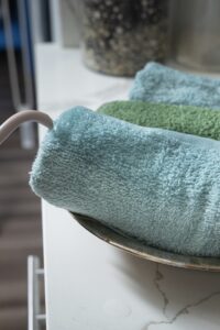 Boiling hot water being poured from a steam kettle onto 3 rolled up cotton towels, saturating them completely. The towels are nestled in a stainless steel pie pan.