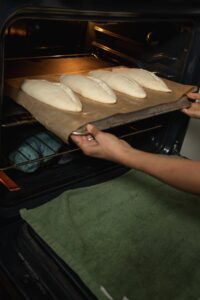 A person using an inverted baking tray as a baking peel to offload four loaves of bread dough into an oven. A green towel is visible on the open oven door.