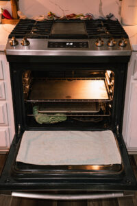 An open oven with a baking steel on the top rack and a baking sheet on the bottom rack. There is a towel draped over the oven door. The vents behind the stovetop are blocked with damp tea towels. Lava rocks and steam towels are placed on the bottom the the oven, below the baking sheet.