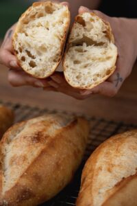 A person holding a freshly baked loaf of birote salado split in half, revealing a fluffy interior. Below, more loaves are cooling on a wire rack. The bread has a golden, crusty exterior.