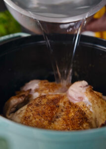 A whole, seasoned chicken is placed in a large pot. Water is being poured into the pot, covering the chicken. The chicken appears to have a golden-brown crust.