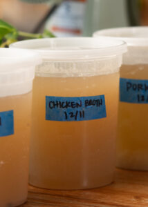 Close-up of containers filled with broth. The one in the foreground is labeled Chicken Broth 12/11 on a blue tape. Other containers are partially visible in the background, including one labeled Pork. The containers are on a wooden surface.