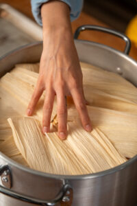 A hand touches corn husks layered on top of a pot, preparing to steam tamales.