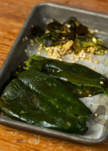 Roasted poblano peppers with some seeds removed, resting on a wooden surface. The deep green peppers glisten slightly, suggesting they are freshly roasted and skinned.