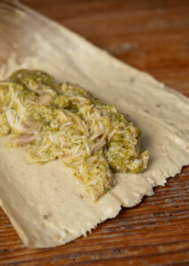 Close-up of a corn husk with a layer of masa and a filling of shredded chicken in green sauce, ready to be wrapped into a tamale, on a wooden surface.