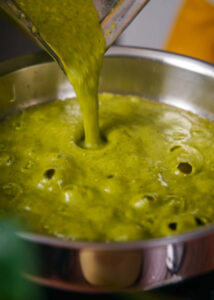 A vibrant green salsa is being poured from a blender into a stainless steel pan. The sauce appears thick and smooth, with bubbles forming as it fries in rendered pork fat.