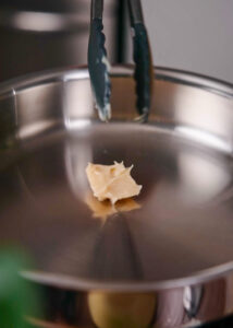 A small dollop of brown rendered pork fat starts to melt in a stainless steel frying pan.