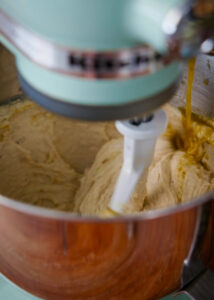 A close-up of a stand mixer in action, with a paddle attachment mixing a fluffy, creamy masa in a metal bowl. A stream of salsa verde and chicken broth are being poured into the mixture from the side.