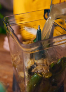 A person pours broth into a blender filled with roasted green peppers, cilantro, and onions.