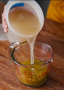 A person pours chicken broth into a glass measuring cup containing a green salsa