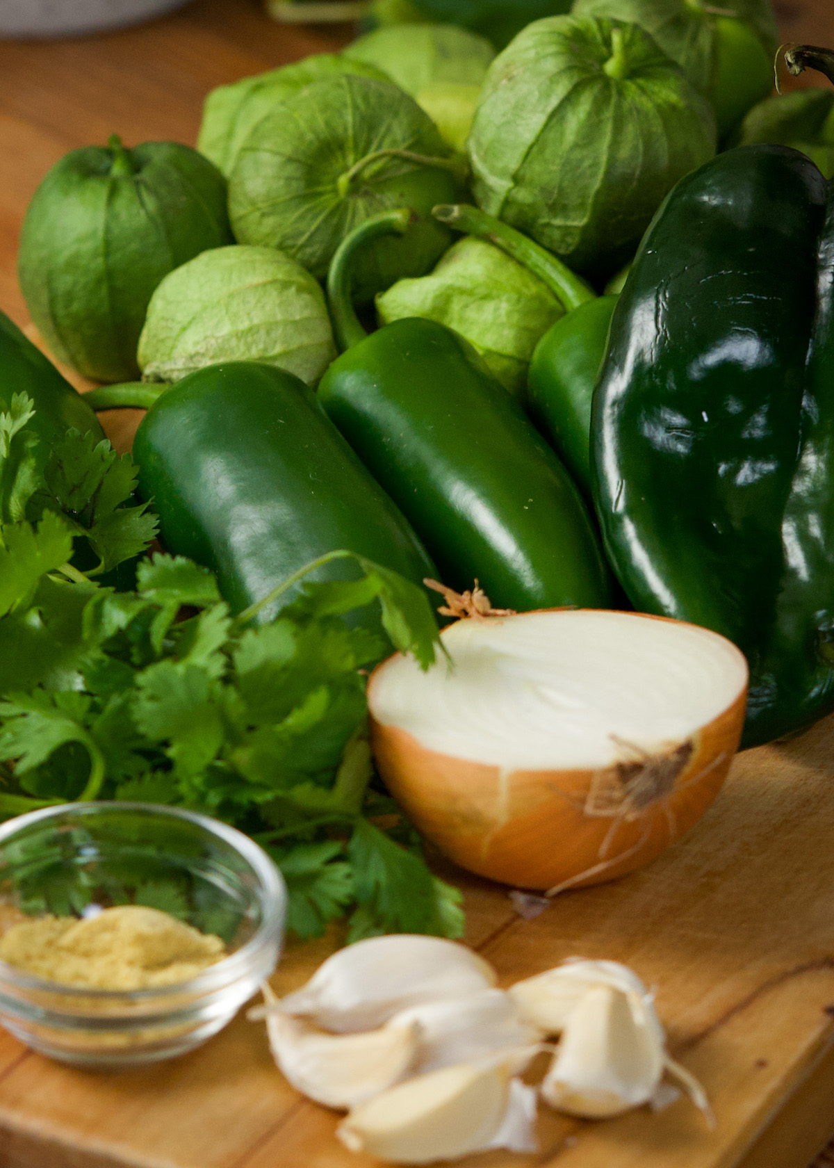 Fresh ingredients on a wooden cutting board include several green tomatillos, jalapeños, a poblano pepper, a halved onion, fresh cilantro, garlic cloves, and a small bowl of seasonings.