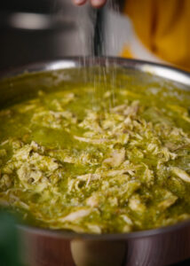 A person sprinkles salt over a pot filled with shredded chicken in a green salsa