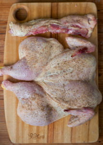 Raw spatchcocked chicken and spine, seasoned with salt & pepper, and a separate leg displayed on a wooden cutting board.