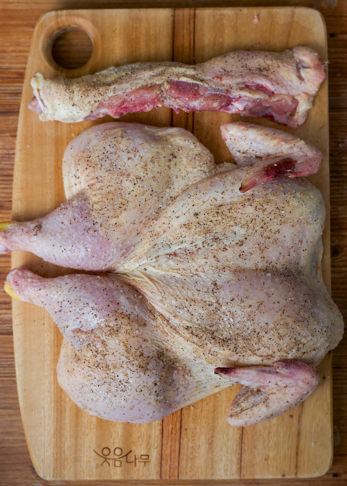 Raw spatchcocked chicken and spine, seasoned with salt & pepper, and a separate leg displayed on a wooden cutting board.