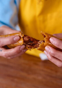 Two hands breaking apart a tamale, revealing its shredded beef in red chile sauce filling. The masa is very moist, with a beef to masa ratio of about 2:1