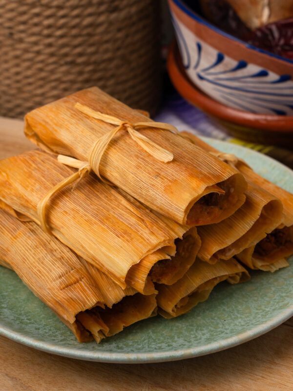 A stack of tamales wrapped in corn husks sits on a green plate. The background includes a rustic rope and a colorful bowl, adding to the traditional setting.