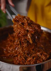 Close-up of a person using tongs to lift a portion of tender, red saucy beef filling for tamales.