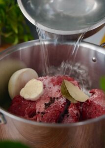 Raw beef, a whole onion, garlic, black peppercorns, salt, and bay leaves are placed in a pot. Cool water is being poured into the pot from a bowl, preparing ingredients for cooking.