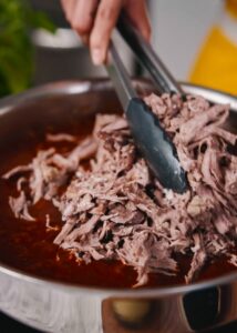 Tongs adding shredded beef into a sautepan of red chili sauce