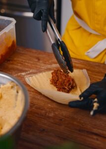 A person wearing gloves uses tongs to fill a corn husk with meat filling on fresh masa.