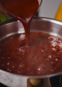 A red chile sauce is being poured from a glass pitcher into a saucepan with lard, creating a steamy and bubbling mixture.