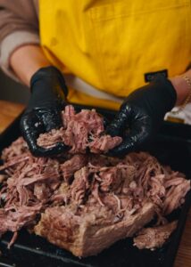 A person wearing a yellow apron and black gloves is shredding slow-cooked beef on a black tray.