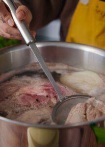 A person uses a skimmer to remove scum from a large pot of simmering broth containing beef , garlic, seasonings, and onions.