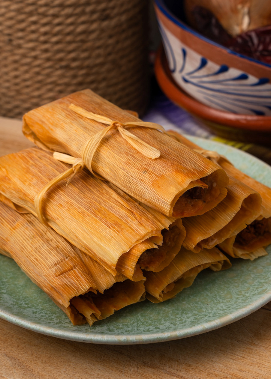 A stack of tamales wrapped in corn husks sits on a green plate. The background includes a rustic rope and a colorful bowl, adding to the traditional setting.